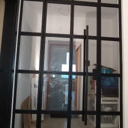 SMART METAL Tomasz Krause - Szklana ściana działowa z czarną metalową ramą, widok na wnętrze domu, częściowo zasłonięte folią zabezpieczającą elementy wyposażenia.