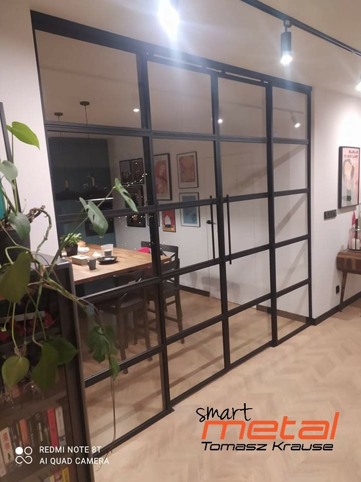 Szklana ściana działowa z czarnymi metalowymi szprosami oddzielająca jadalnię, widoczny stół z nakryciem, w tle oświetlenie szynowe
