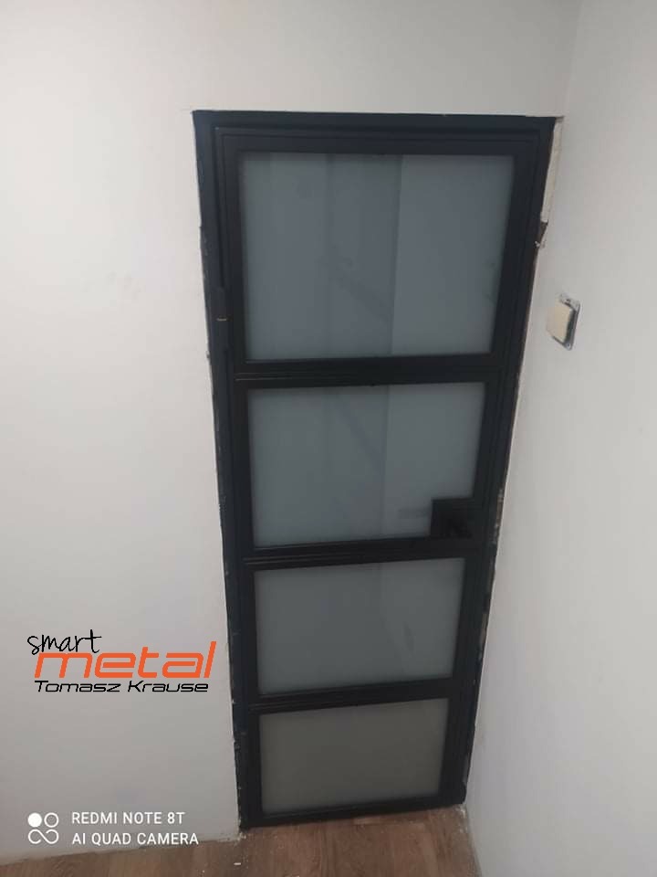 Czarna, metalowa konstrukcja drzwi z trzema szklanymi panelami, widoczna w białym pomieszczeniu, z logo firmy Smart Metal w lewym dolnym rogu.
