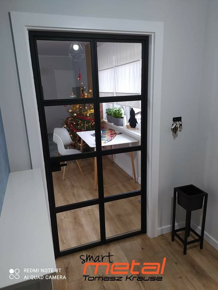 Szklane drzwi wewnętrzne z czarną, metalową ramą w stylu loft, widok na świątecznie udekorowane wnętrze z choinką, jasne panele podłogowe i białe ściany.