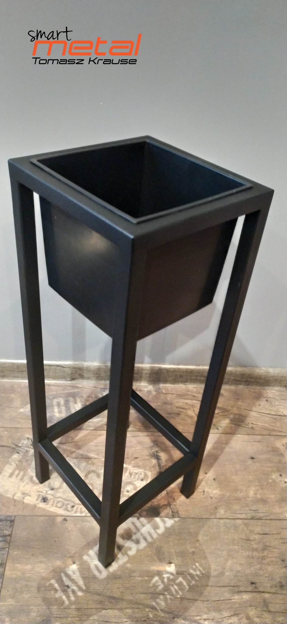 Czarna, metalowa konstrukcja kwietnika z logo firmy Smart Metal Tomasz Krause w tle, ustawiona na podłodze z wzorem imitującym stare napisy.