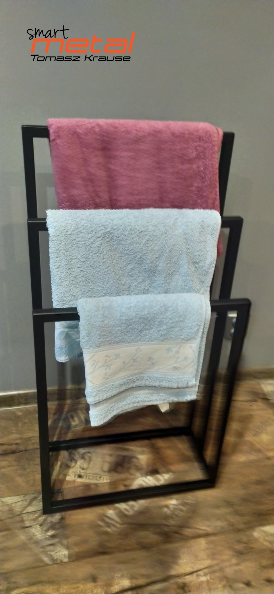 Czarny metalowy stojak na ręczniki z trzema poziomami, na których wiszą różowe i błękitne ręczniki, widoczne logo firmy w tle.