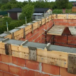 Wznoszenie murów z czerwonej cegły z drewnianym szalunkiem wypełnionym świeżym betonem, widok z góry na budowę domu w otoczeniu zieleni.