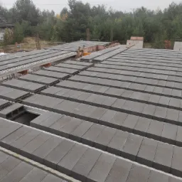 Widok z góry na budowę stropu domu z szarych, betonowych elementów, z widocznym zbrojeniem i otworami, na tle jesiennego lasu i częściowo wykończonego budynku.