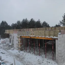 Wznoszenie ścian fundamentowych z bloczków betonowych w zimowej scenerii, widoczne szalunki i podpory, w tle las i przyczepa firmy budowlanej.