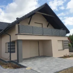 Nowoczesny dom z szarą, poziomą balustradą na balkonie, widok z dołu na elewację i podjazd z kostki brukowej w szarym odcieniu.