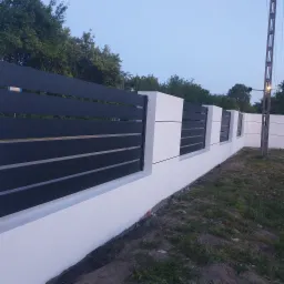 Nowoczesne ogrodzenie z poziomych, czarnych paneli zamocowanych między białymi, betonowymi słupkami, widoczne na tle zieleni i błękitnego nieba o zmierzchu.