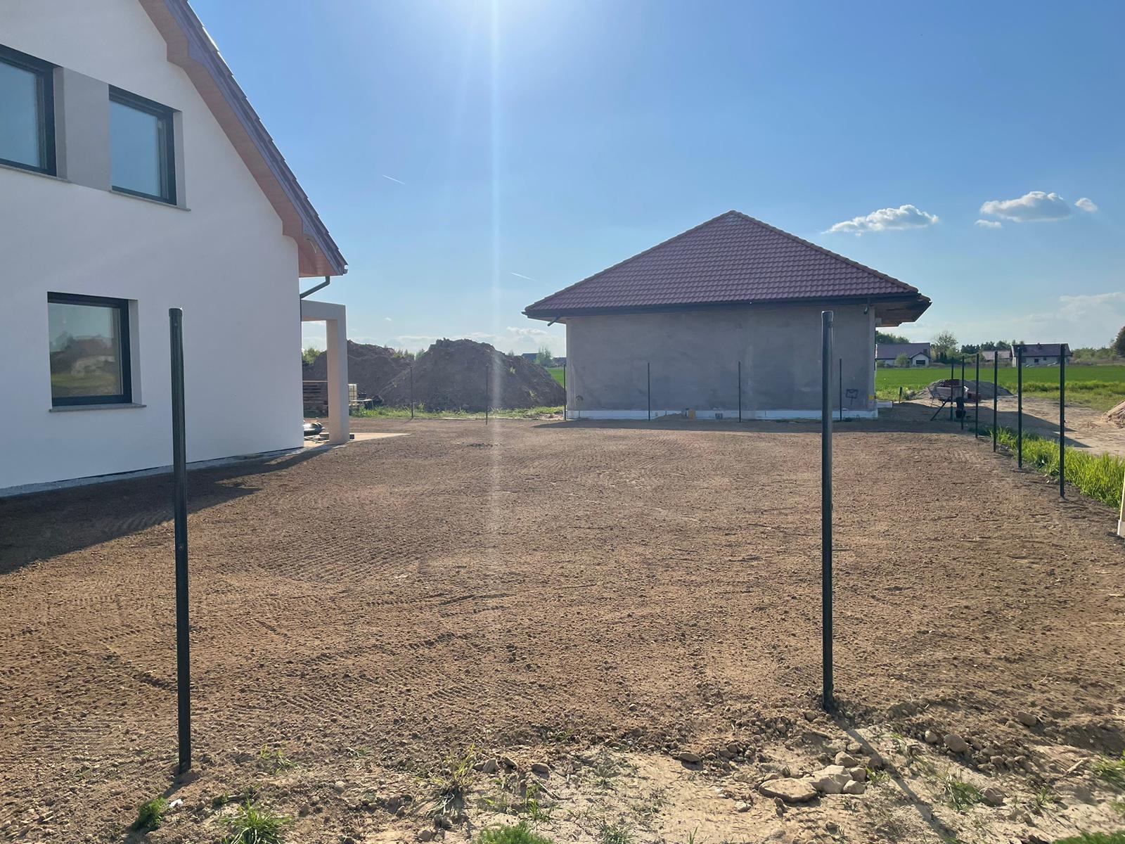 Przygotowany, wyrównany teren pod trawnik, widoczne ślady po maszynach, w tle nowo wybudowany dom i budynek gospodarczy, częściowo ogrodzony. Słoneczny dzień.