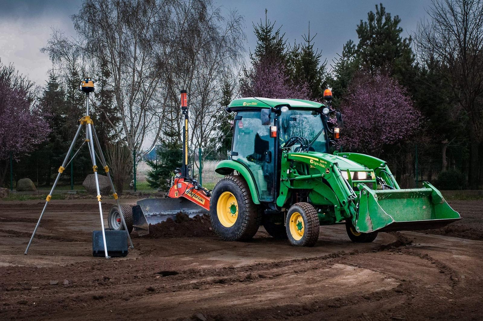 Zielony traktor John Deere z żółtymi felgami wyrównuje teren pod przyszły trawnik, obok niwelator laserowy na statywie, w tle drzewa z delikatnymi różowymi kwiatami.