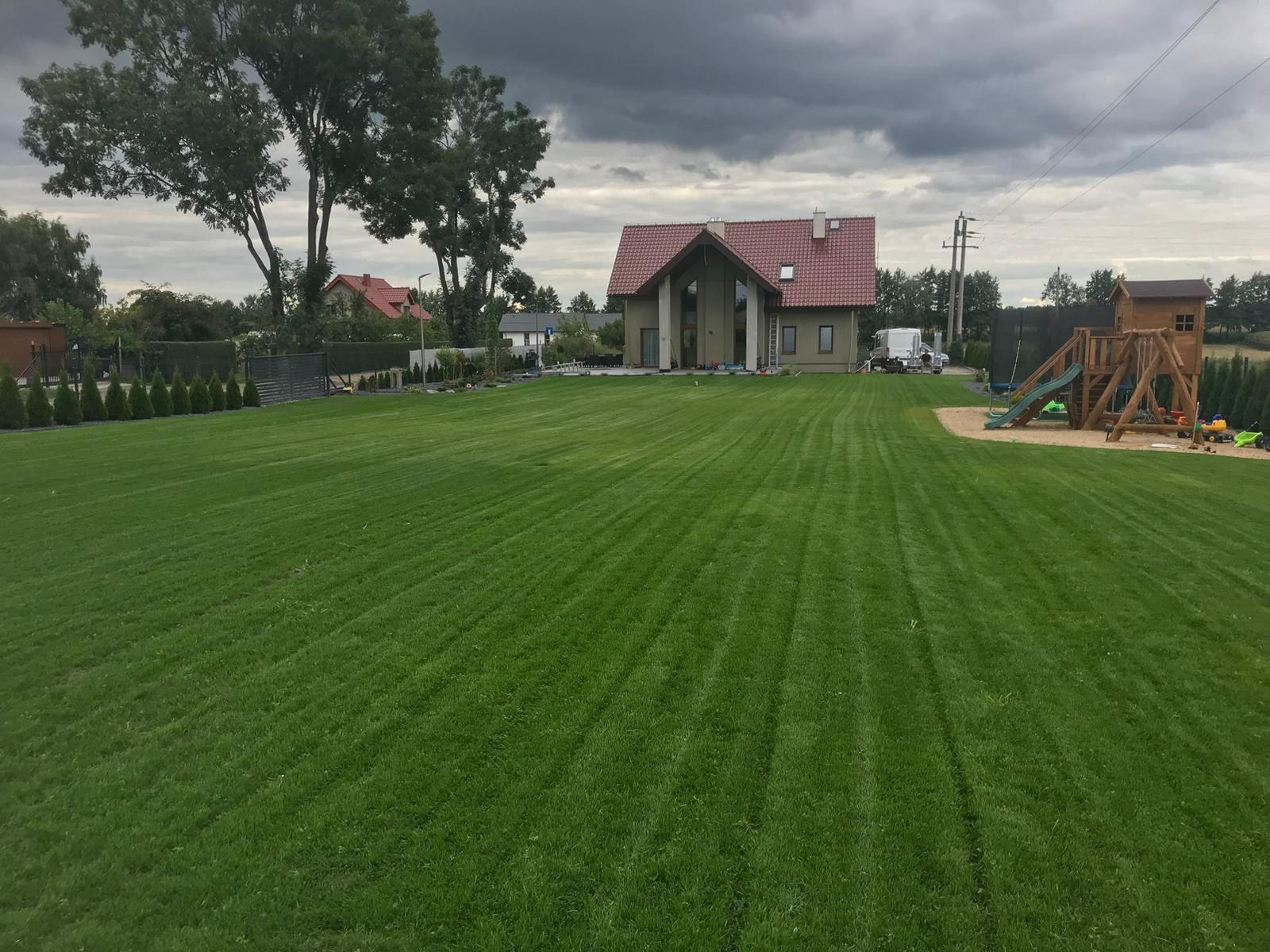 Rozległy, intensywnie zielony trawnik przed nowoczesnym domem z czerwonym dachem, widoczne ślady po koszeniu, w tle plac zabaw dla dzieci i ciemne, burzowe niebo.