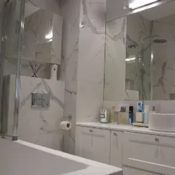 Nowoczesna łazienka z białymi marmurowymi płytkami, kabiną prysznicową i wanną, białą szafką pod umywalką z chromowanymi uchwytami oraz dekoracyjnymi flakonami na blacie.