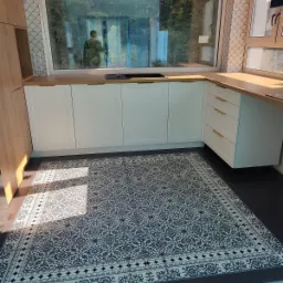 Jasna kuchnia z białymi szafkami, drewnianym blatem i wzorzystą podłogą w stylu vintage, widok na zewnątrz przez duże okno.