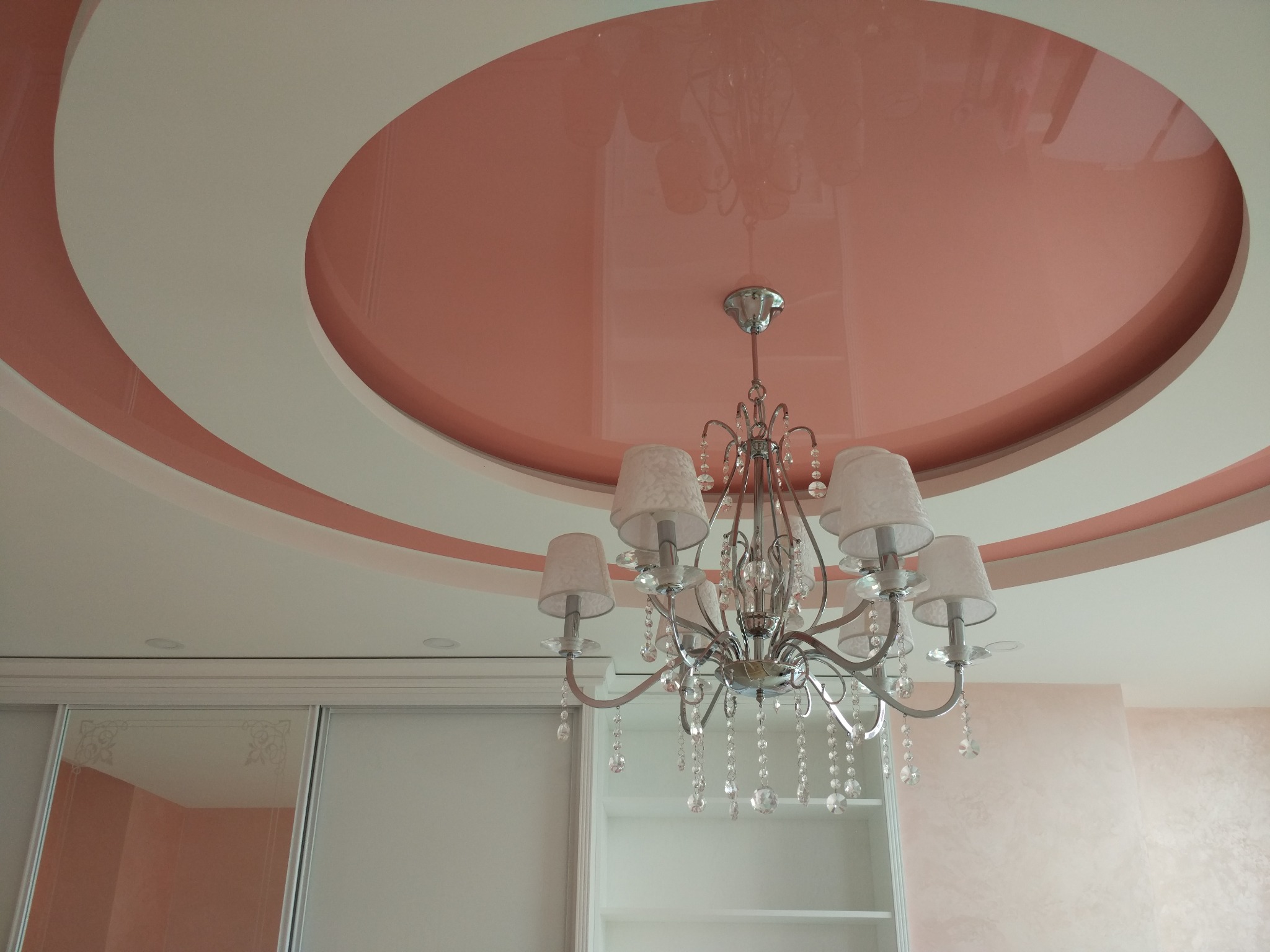 Sufit podwieszany z okrągłym, lakierowanym elementem w kolorze łososiowym i ozdobnym żyrandolem z kryształkami, widoczna szafa przesuwna z lustrami.