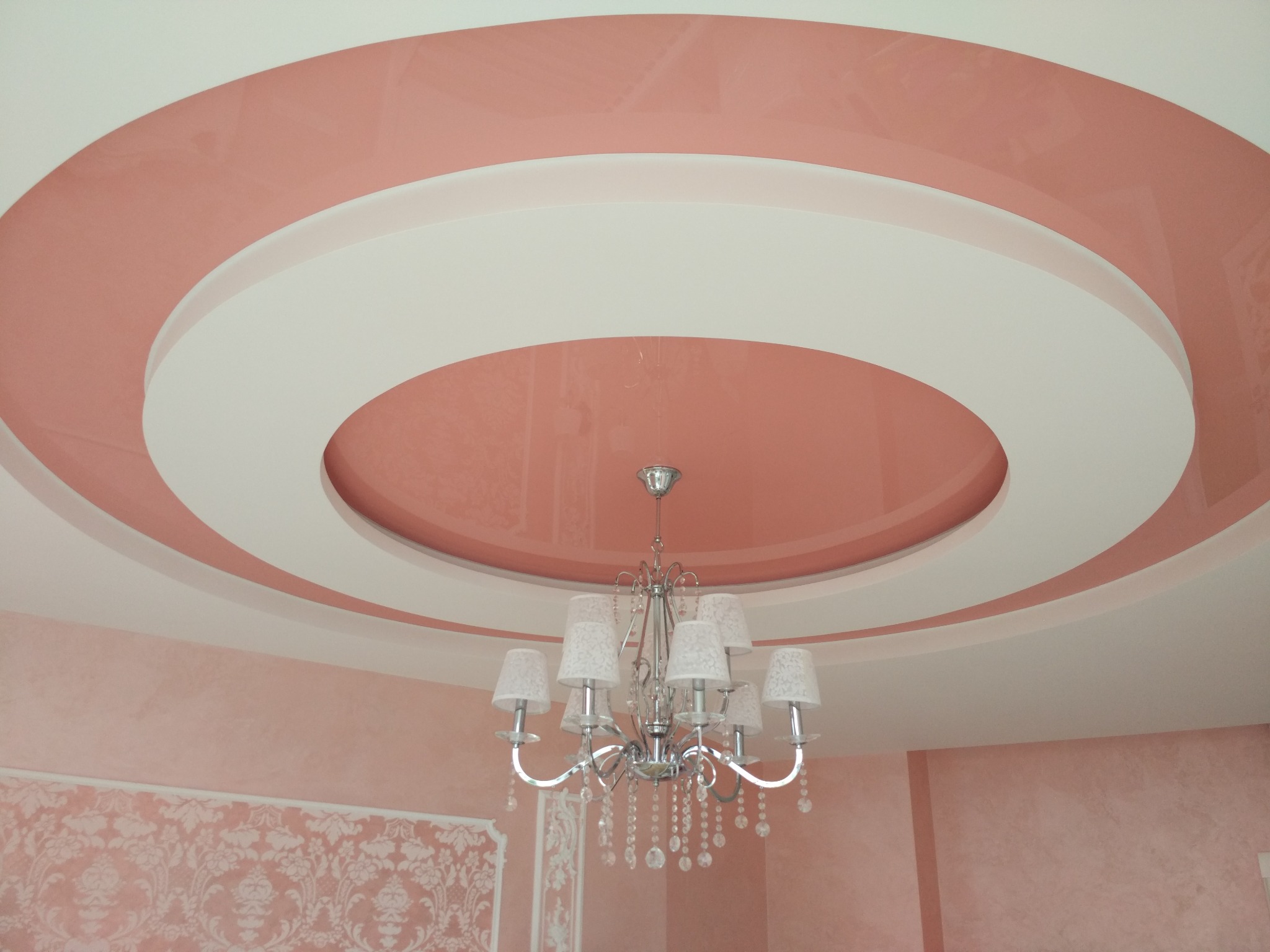 Różowy, lakierowany sufit podwieszany z okrągłymi wnękami i białym obramowaniem, ozdobiony klasycznym, srebrnym żyrandolem z białymi abażurami i kryształkami, ściana w tle z dekoracyjnym wzorem.