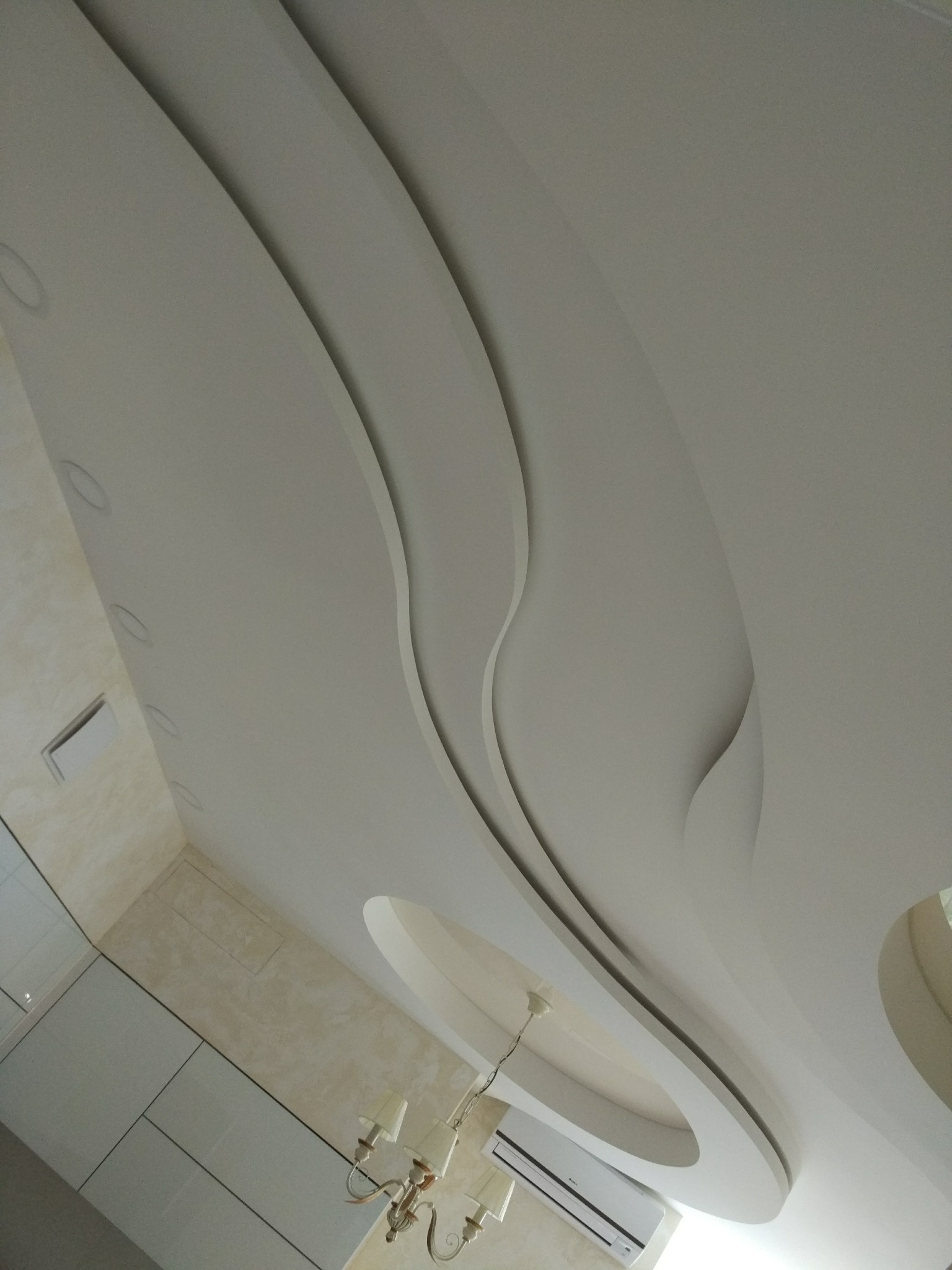 Nowoczesny sufit podwieszany z dekoracyjnymi, falistymi wgłębieniami i oświetleniem punktowym, uzupełniony o klasyczny żyrandol i klimatyzator ścienny.