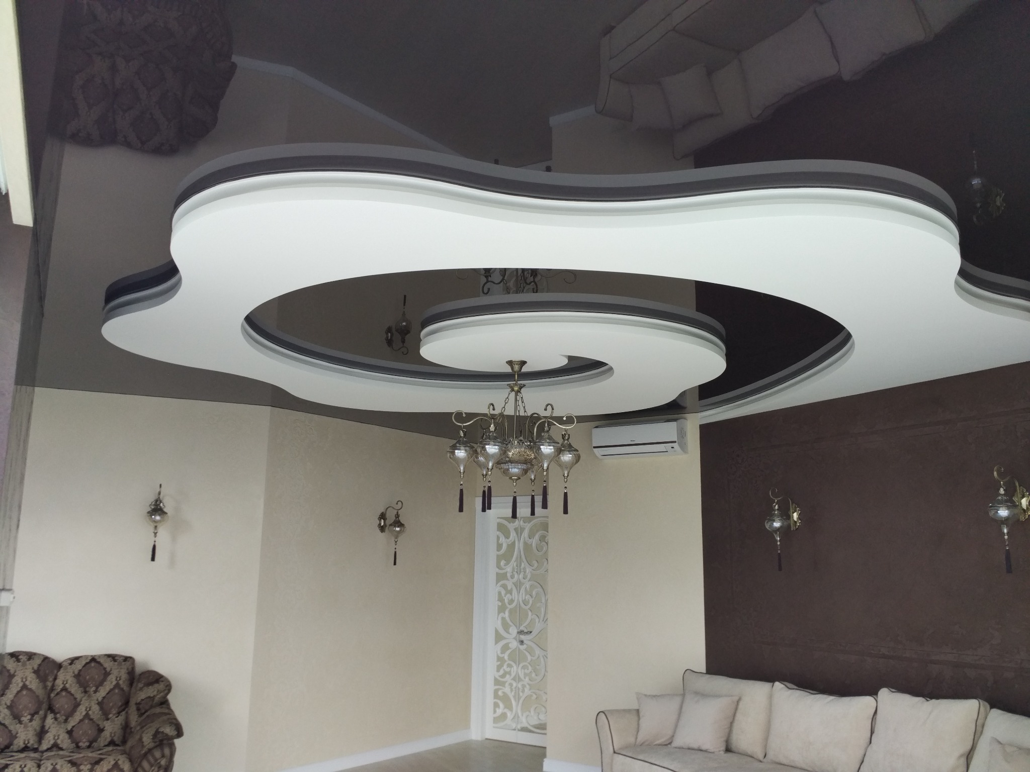 Nowoczesny salon z dekoracyjnym, podwieszanym sufitem o spiralnym kształcie, oświetlony kryształowym żyrandolem i kinkietami, z klimatyzacją na ścianie.