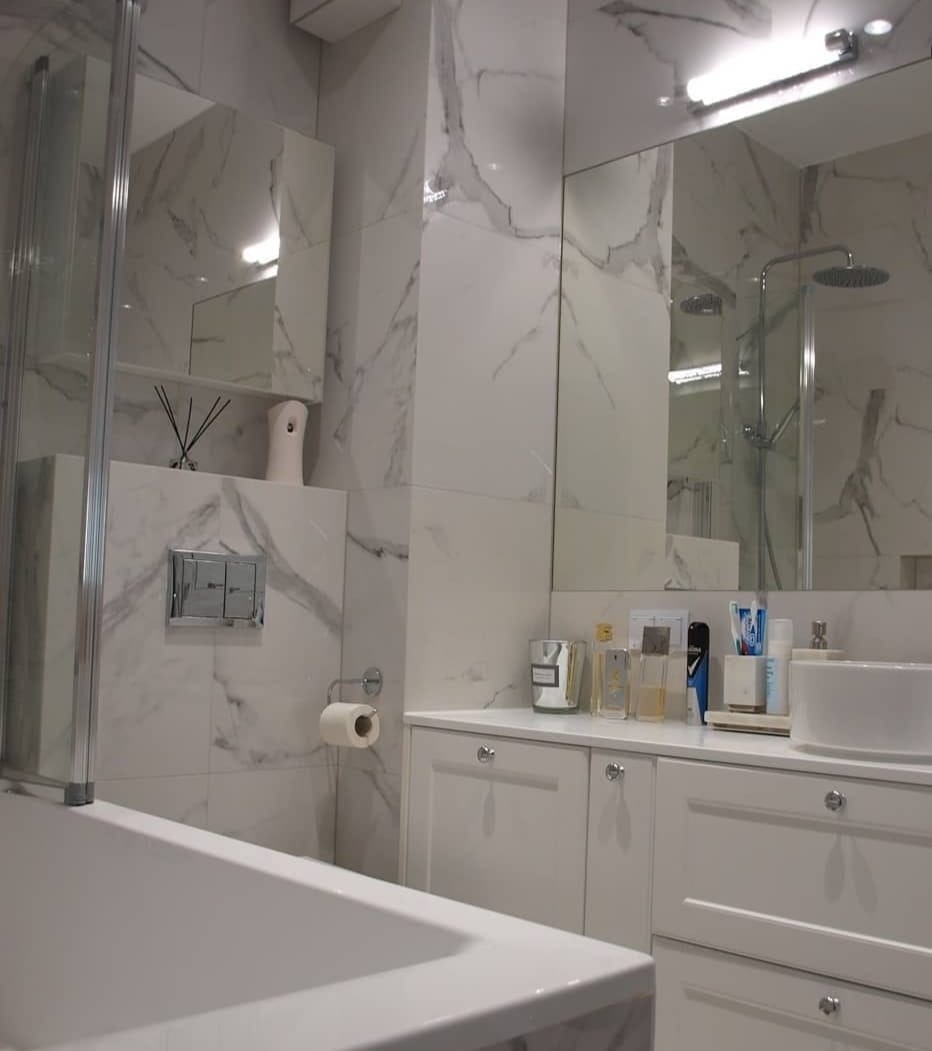Nowoczesna łazienka z białymi marmurowymi płytkami, kabiną prysznicową i wanną, białą szafką pod umywalką z chromowanymi uchwytami oraz dekoracyjnymi flakonami na blacie.