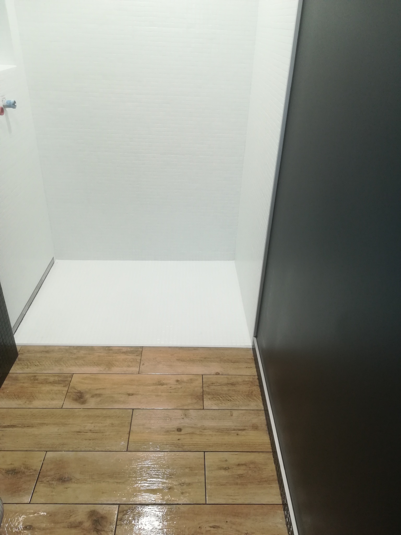 Nowoczesna kabina prysznicowa z drewnopodobnymi płytkami podłogowymi, białymi ścianami wyłożonymi drobną mozaiką i ciemną ścianą boczną.