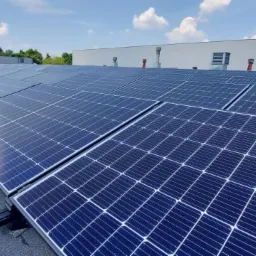 Rząd paneli słonecznych na płaskim dachu z widoczną ceglaną konstrukcją kominową w tle i błękitnym niebem z lekkimi chmurami.