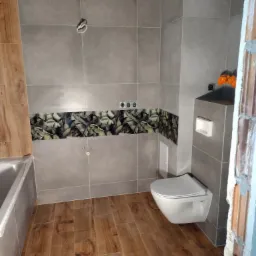 Szare płytki w łazience z dekoracyjnym pasem z motywem liści, drewnopodobna podłoga i podwieszana toaleta w trakcie montażu.