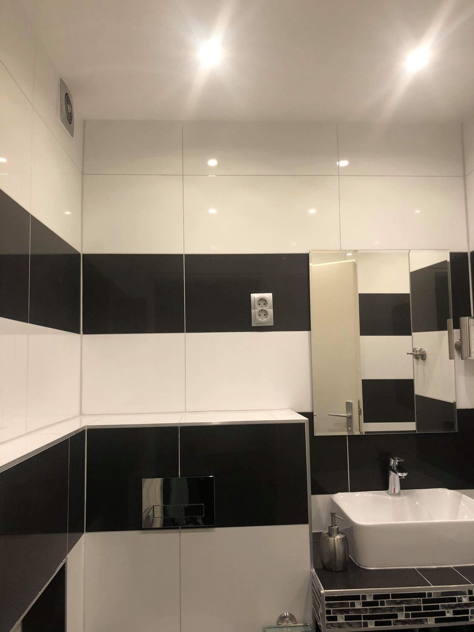 Nowoczesna łazienka z czarno-białymi, poziomymi pasami płytek, kwadratową umywalką i lustrem z drzwiczkami.