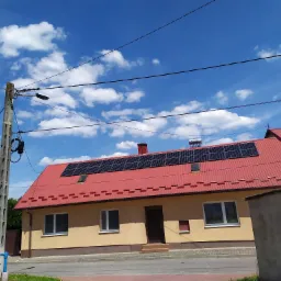 Dom jednorodzinny z czerwoną dachówką i panelami fotowoltaicznymi na dachu, widok z ulicy w słoneczny dzień z błękitnym niebem i chmurami.