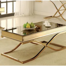 Stolik kawowy z lustrzanym blatem i złotą, krzyżową podstawą, na blacie owoce i filiżanki, w tle okno z białymi zasłonami.