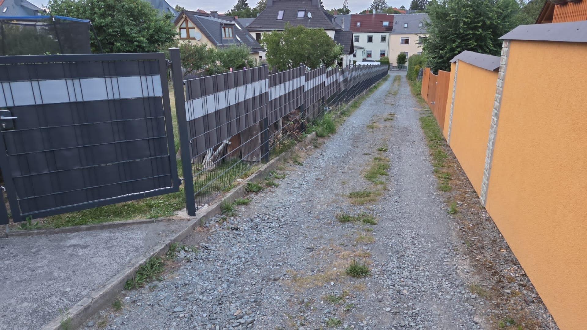 Otwarta brama panelowa z szarego metalu, w tle długa linia ogrodzenia panelowego z poziomymi, jasnymi pasami. Widok na żwirową drogę między ogrodzeniami.