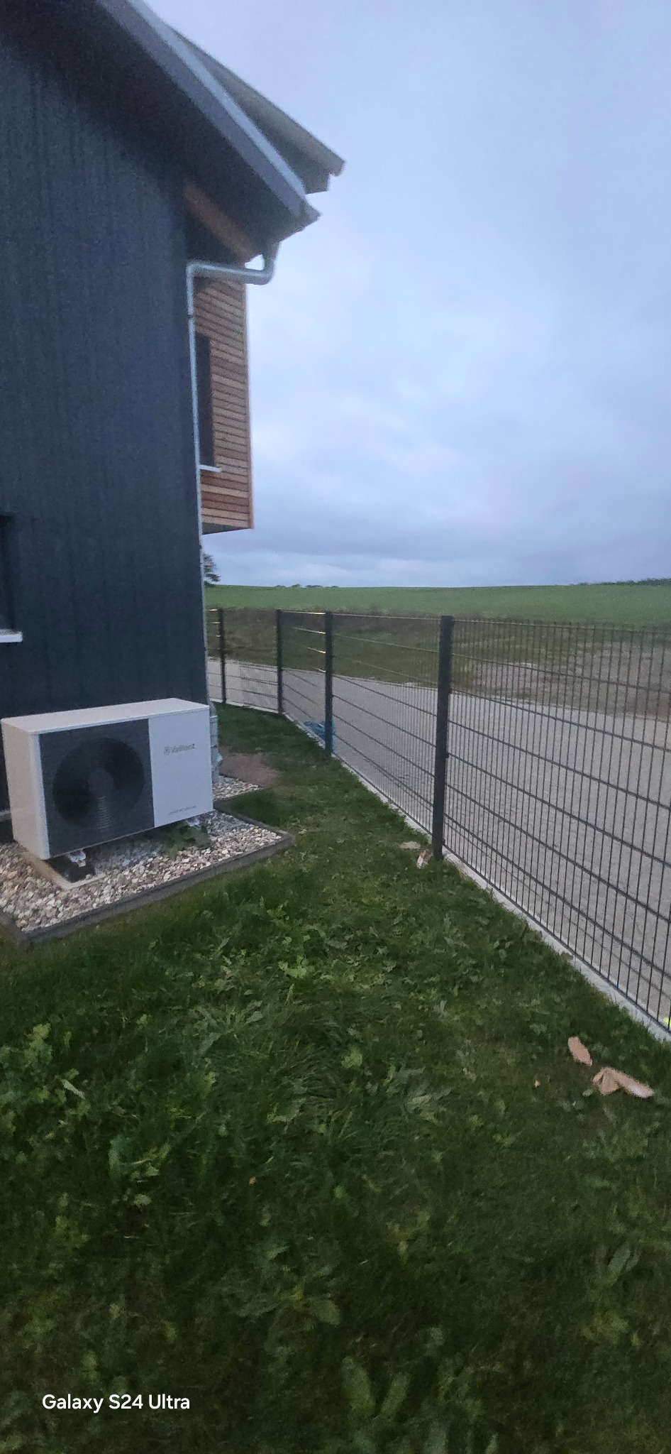 Nowoczesne ogrodzenie panelowe w kolorze grafitowym, zamontowane przy posesji z pompą ciepła Vaillant. Widok na zielone pole w pochmurny dzień.