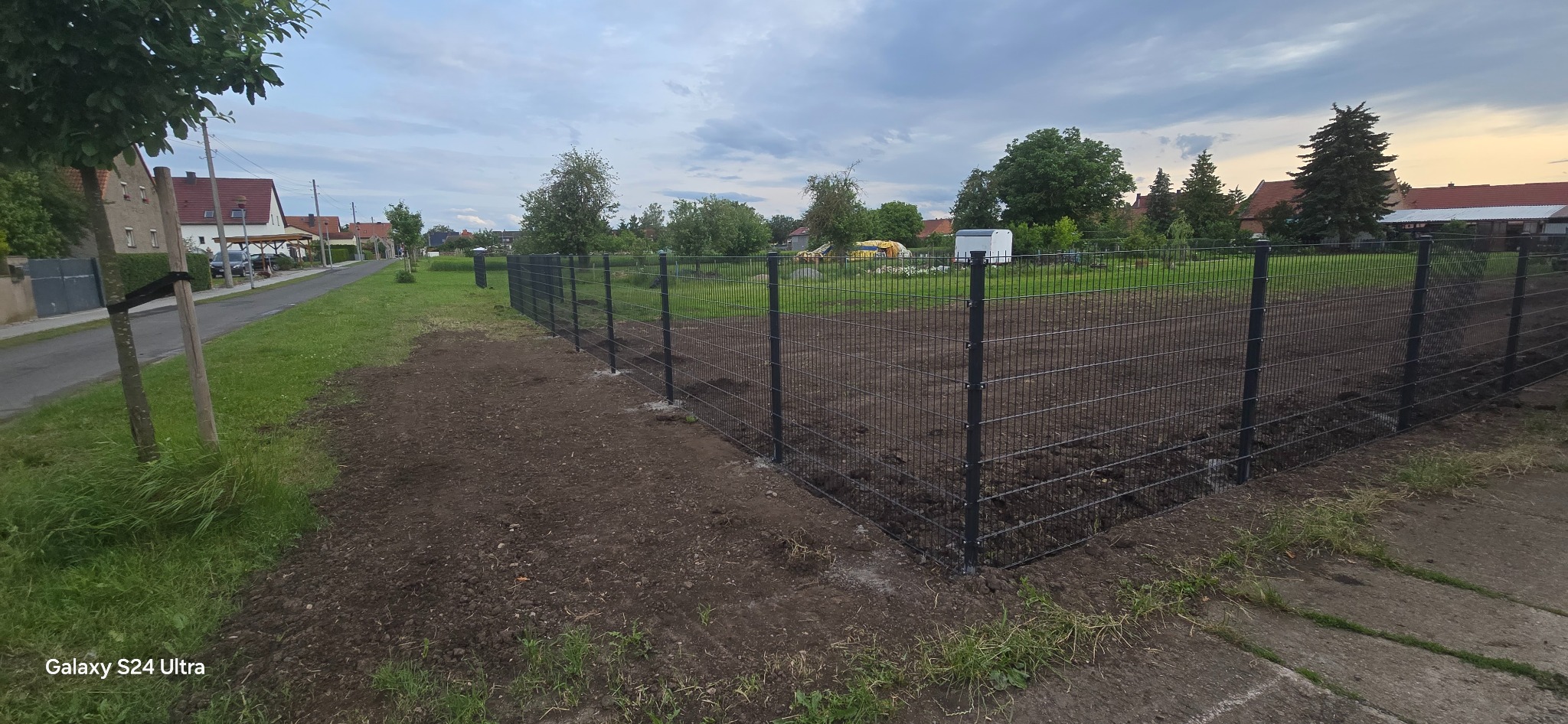 Nowoczesne, grafitowe ogrodzenie panelowe z podmurówką na świeżo wyrównanym terenie działki, widok z zewnątrz. Tło: zabudowa jednorodzinna, pochmurne niebo.