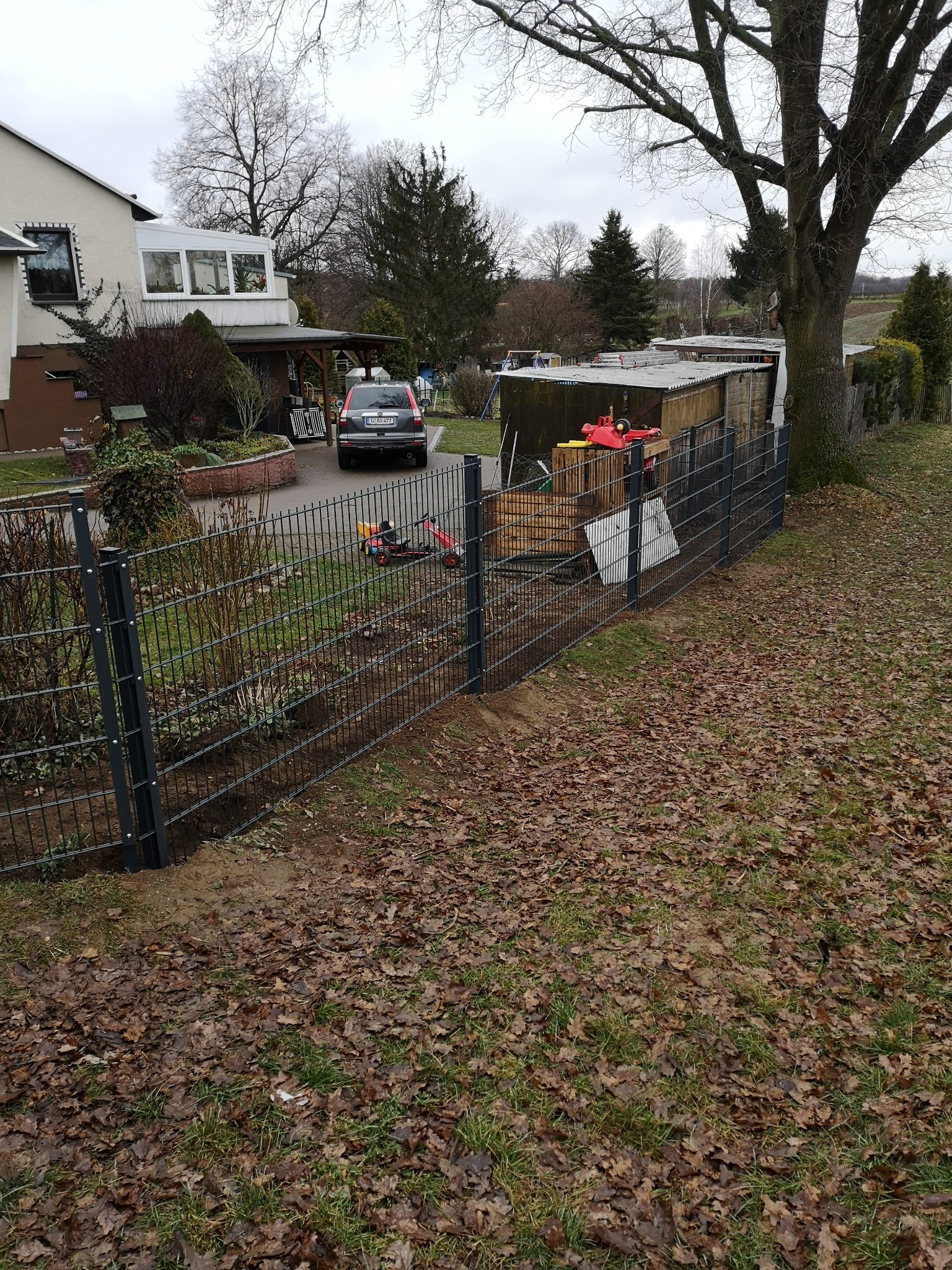 Częściowo zamontowane, nowoczesne ogrodzenie panelowe w kolorze grafitowym, widoczne na tle domu jednorodzinnego i trawnika pokrytego liśćmi. Etap prac montażowych.