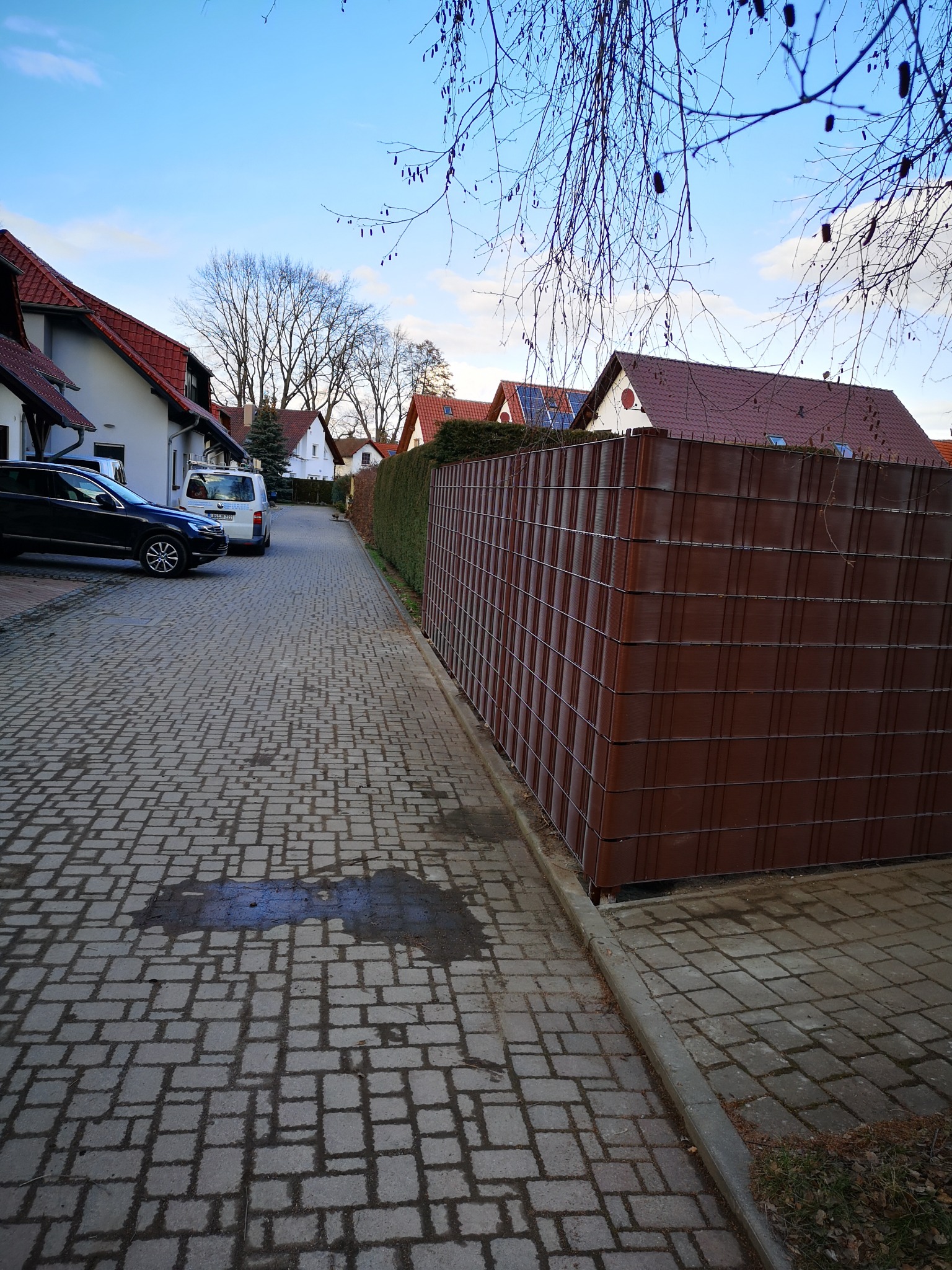 Brązowe ogrodzenie panelowe z metalową siatką na tle domów jednorodzinnych i brukowanej drogi. Widok z perspektywy ulicy w słoneczny dzień.