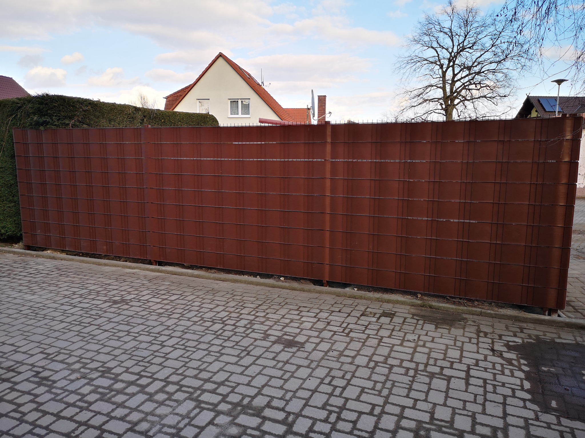 Nowoczesne, brązowe ogrodzenie panelowe z poziomymi prętami, oddzielające posesję od chodnika z kostki. W tle dom z czerwonym dachem i zielony żywopłot.