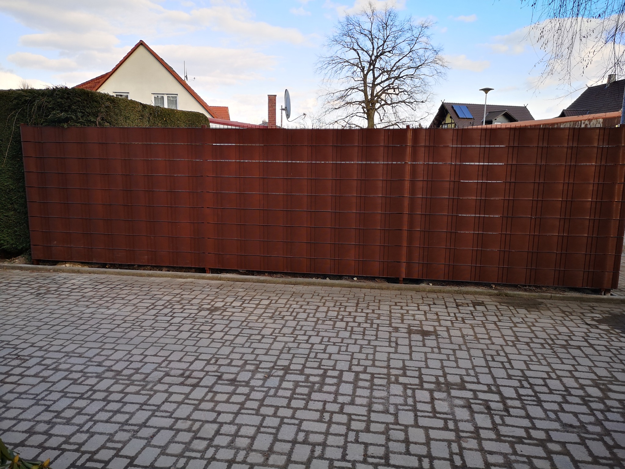 Rustykalne, poziome panele ogrodzeniowe z metalu kortenowskiego, widoczne z poziomu chodnika z kostki brukowej, zieleń w tle i fragmenty domów.