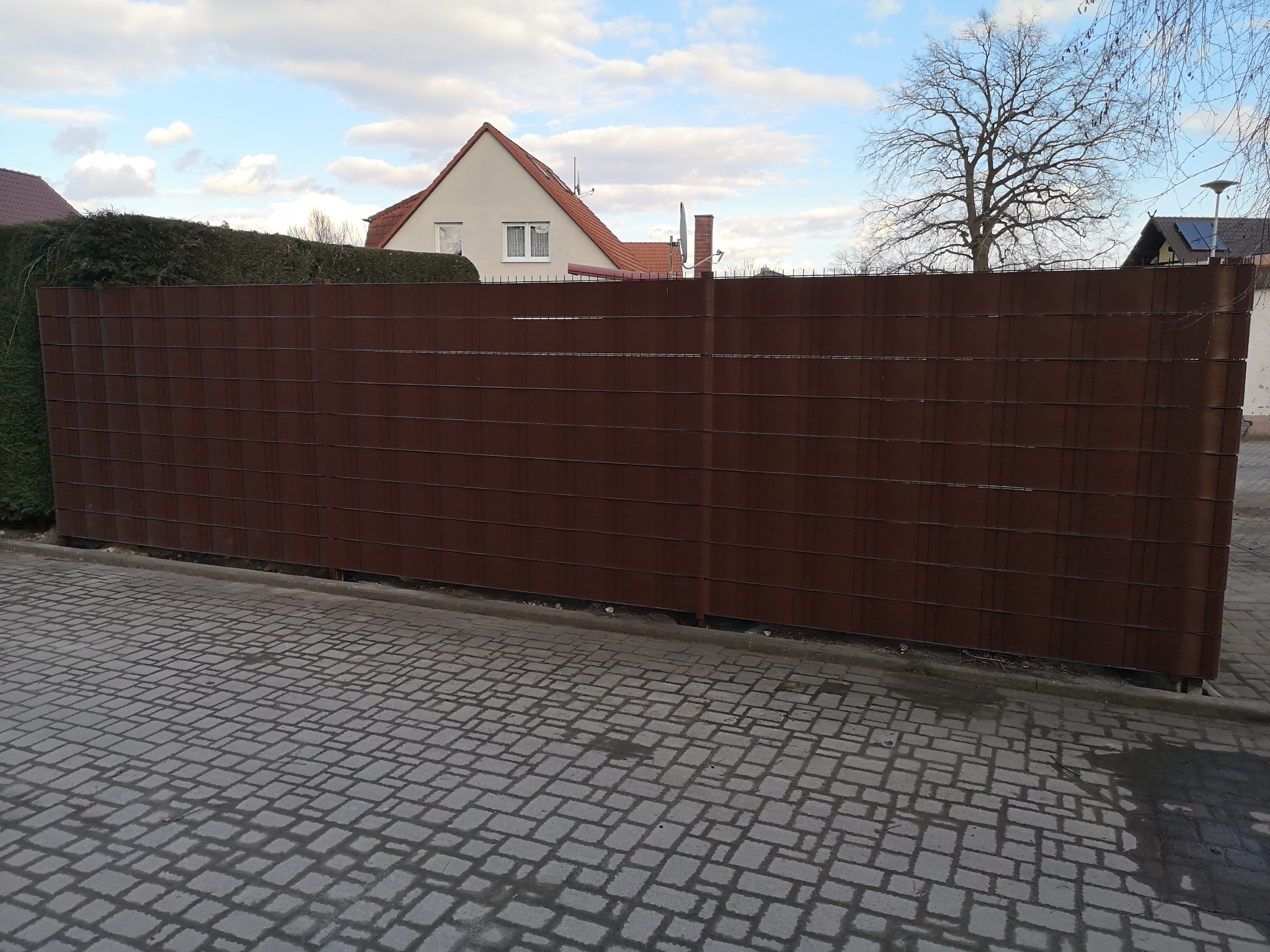 Długi, brązowy panelowy płot z poziomych listew, oddzielający posesję od chodnika z kostki. W tle dom z czerwonym dachem i drzewo bez liści pod błękitnym niebem.