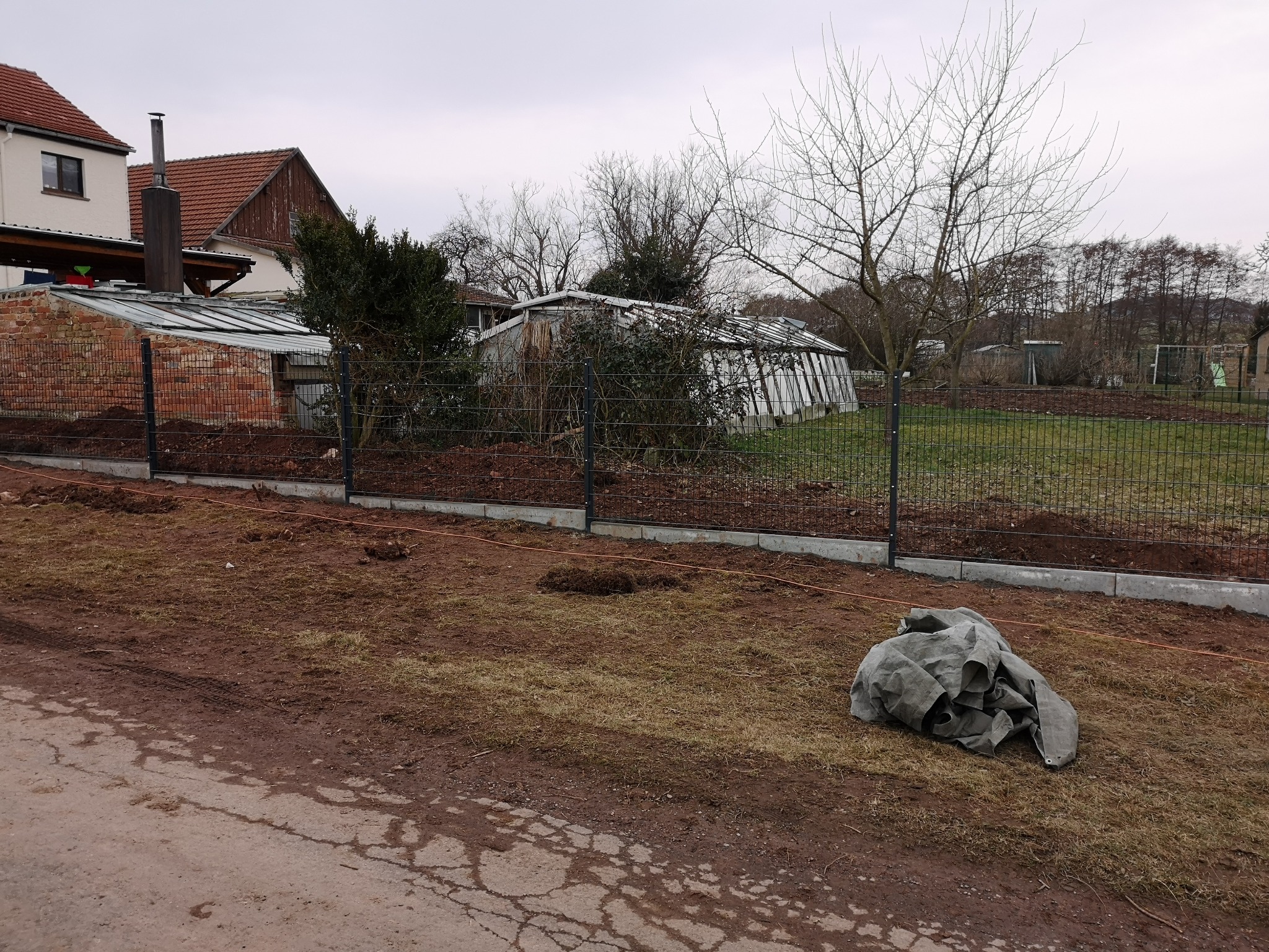 Montaż ogrodzenia panelowego na podmurówce betonowej. Widoczne budynki w tle i rozkopana ziemia. Częściowo ukończony montaż.