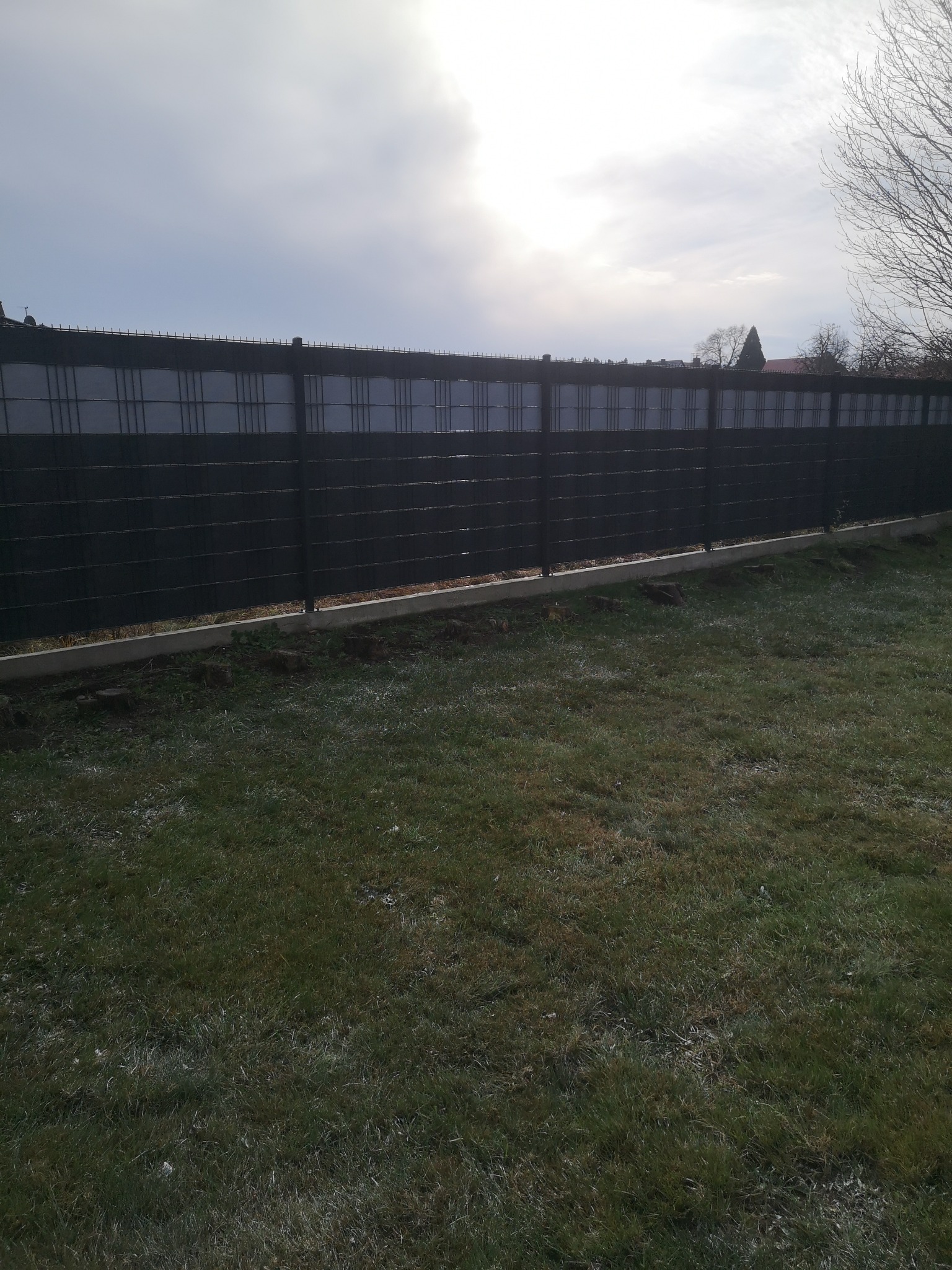 Nowoczesne, czarne ogrodzenie panelowe z podmurówką betonową, widoczne na tle trawnika i pochmurnego nieba. Minimalistyczny design, równomierne panele, świeżo posadzona trawa.