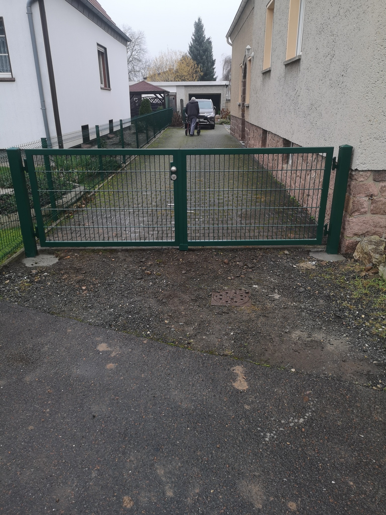 Zielona, metalowa brama dwuskrzydłowa z siatki, zamontowana na wjeździe do posesji. W tle widoczny mężczyzna z wózkiem i zaparkowany samochód.