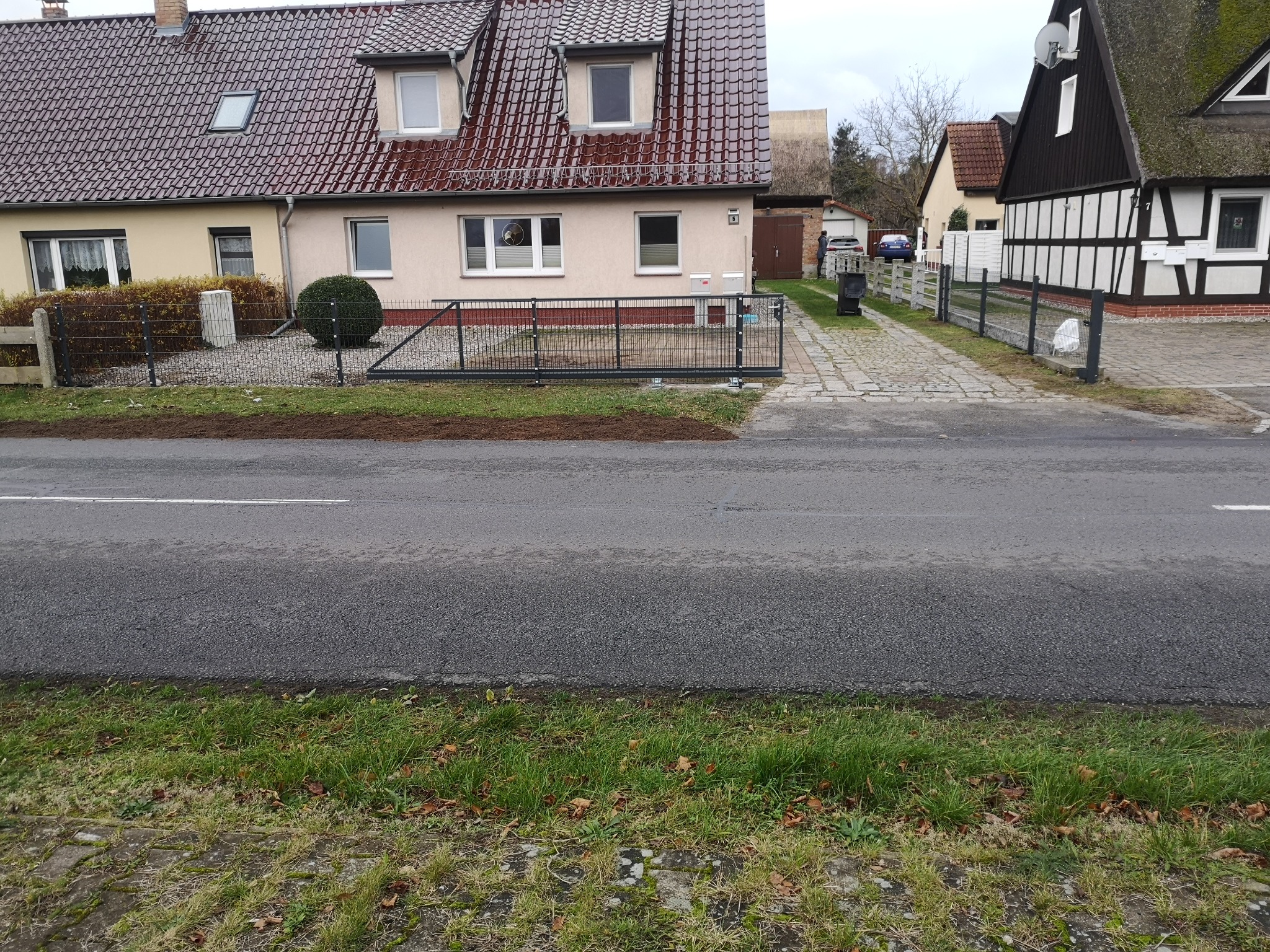 Czarna, przesuwna brama panelowa z siatki na posesji z dwoma domami o różnej architekturze, widok z ulicy. Brama zamontowana na płaskim terenie.