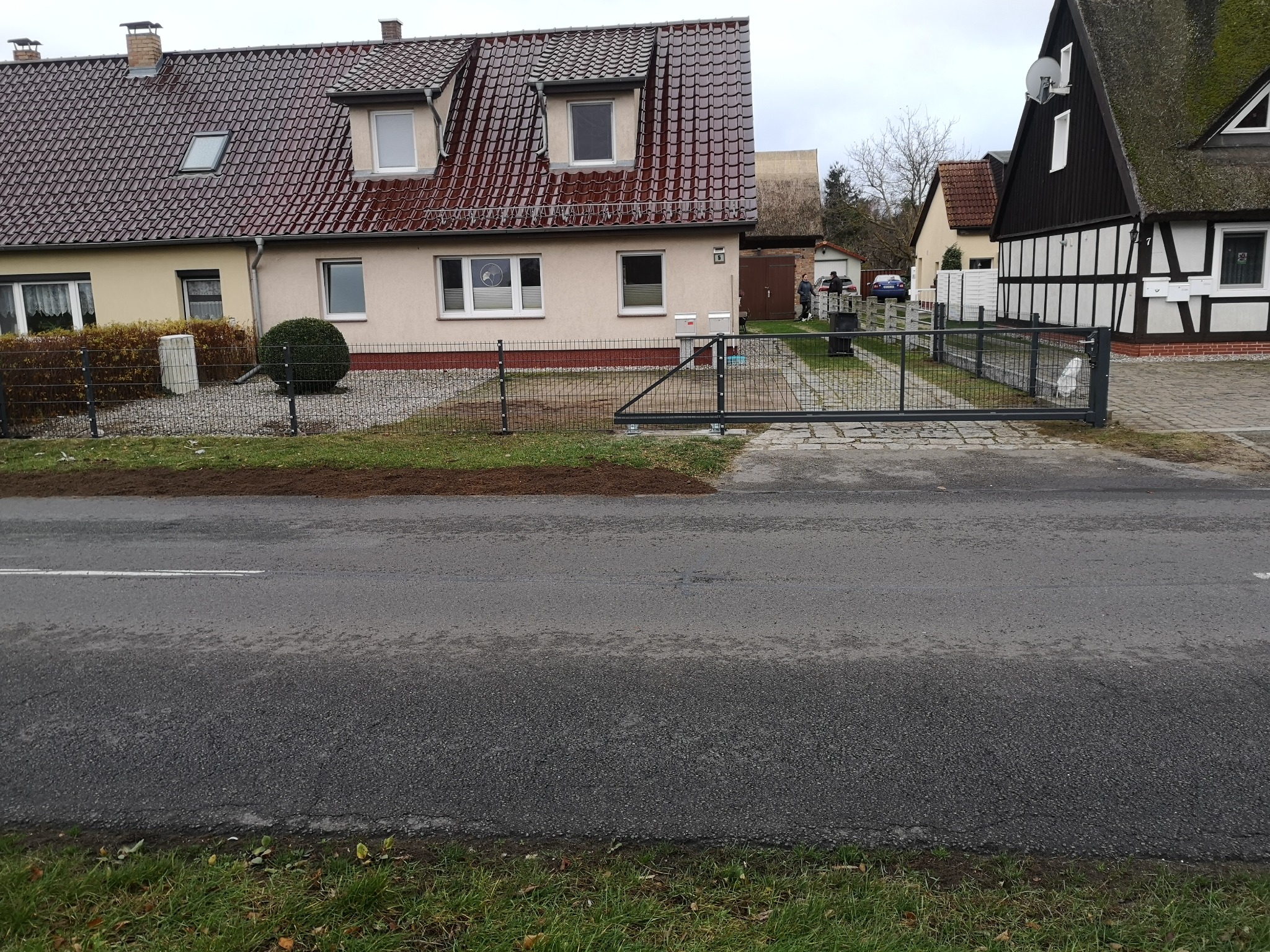 Nowoczesna, przesuwna brama w kolorze grafitowym, zamontowana przy posesji z niskim ogrodzeniem z siatki. Widok z ulicy, dom w tle.