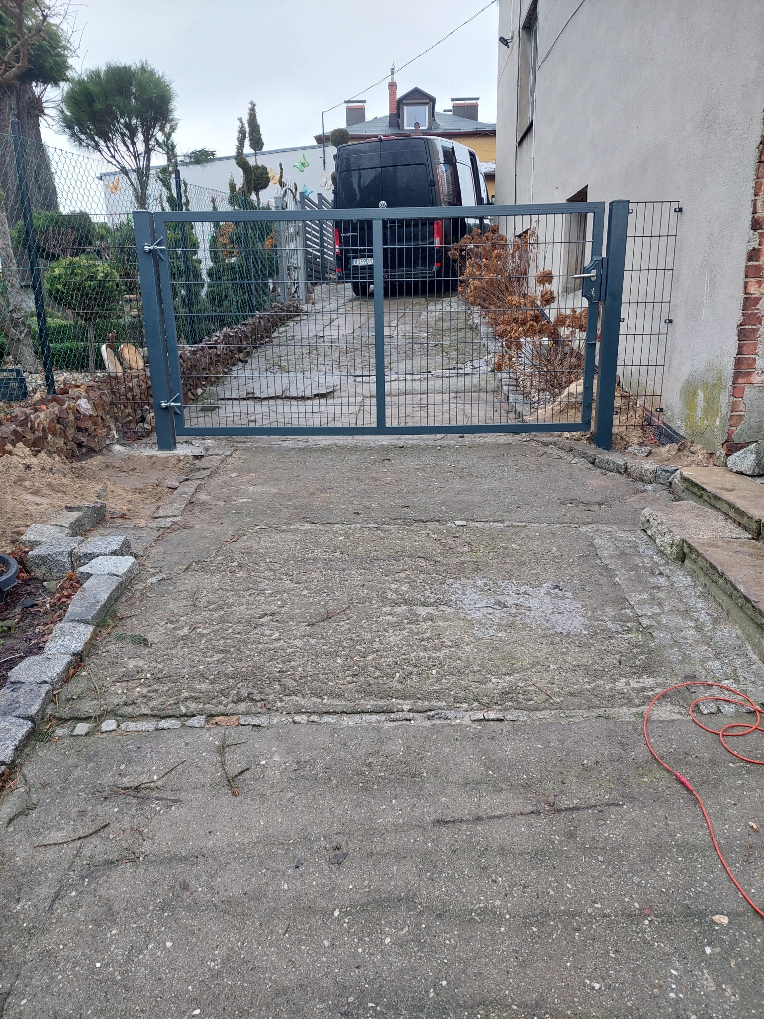 Szara, metalowa brama panelowa z siatki, zamontowana na betonowym podjeździe. W tle zaparkowany czarny van i budynek. Widoczne elementy ogrodu i kostka brukowa.