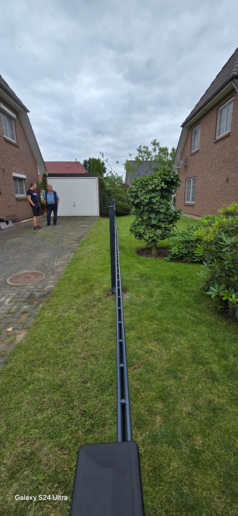 Montaż przęsła ogrodzeniowego na posesji z dwoma mężczyznami w tle. Widok z dołu, na pierwszym planie fragment ogrodzenia, trawnik, budynki mieszkalne.