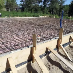 Zbrojenie fundamentu z drewnianym szalunkiem i izolacją termiczną, widoczne czarne rury instalacyjne oraz piasek w wykopie, w tle drzewa.