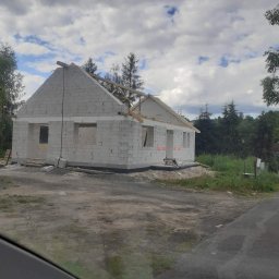 ERDO Polska - Surowy stan budynku w trakcie budowy, ściany wzniesione z białych bloczków, widoczne otwory okienne i drzwiowe, częściowo wykonana konstrukcja dachu, tło stanowią drzewa i pochmurne niebo.