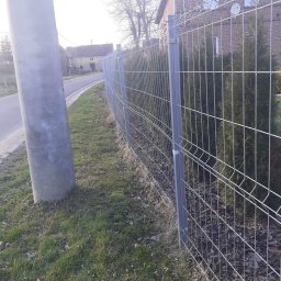 ERDO Polska - Szara, metalowa siatka ogrodzeniowa panelowa zamontowana wzdłuż drogi, z widocznymi słupkami i elementami mocującymi, za ogrodzeniem posadzone tuje, w tle budynek z czerwonej cegły i słup...
