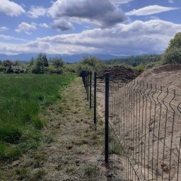 ERDO Polska - Montaż czarnego ogrodzenia panelowego na granicy działki z wysoką trawą i usypanym wzgórzem ziemi w tle, widok na góry pod błękitnym, pochmurnym niebem.