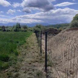 Montaż czarnego ogrodzenia panelowego na granicy działki z wysoką trawą i usypanym wzgórzem ziemi w tle, widok na góry pod błękitnym, pochmurnym niebem.