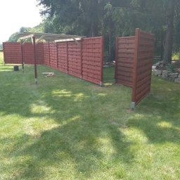 ERDO Polska - Brązowe, drewniane panele ogrodzeniowe z metalowymi słupkami, częściowo połączone drewnianą konstrukcją pergoli, rozstawione na trawniku w ogrodzie.