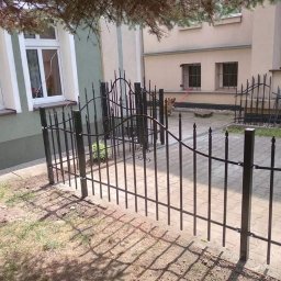 ERDO Polska - Nowoczesne czarne metalowe ogrodzenie posesji z dekoracyjnymi grotami, widok na budynek z oknami i psa na smyczy w tle.