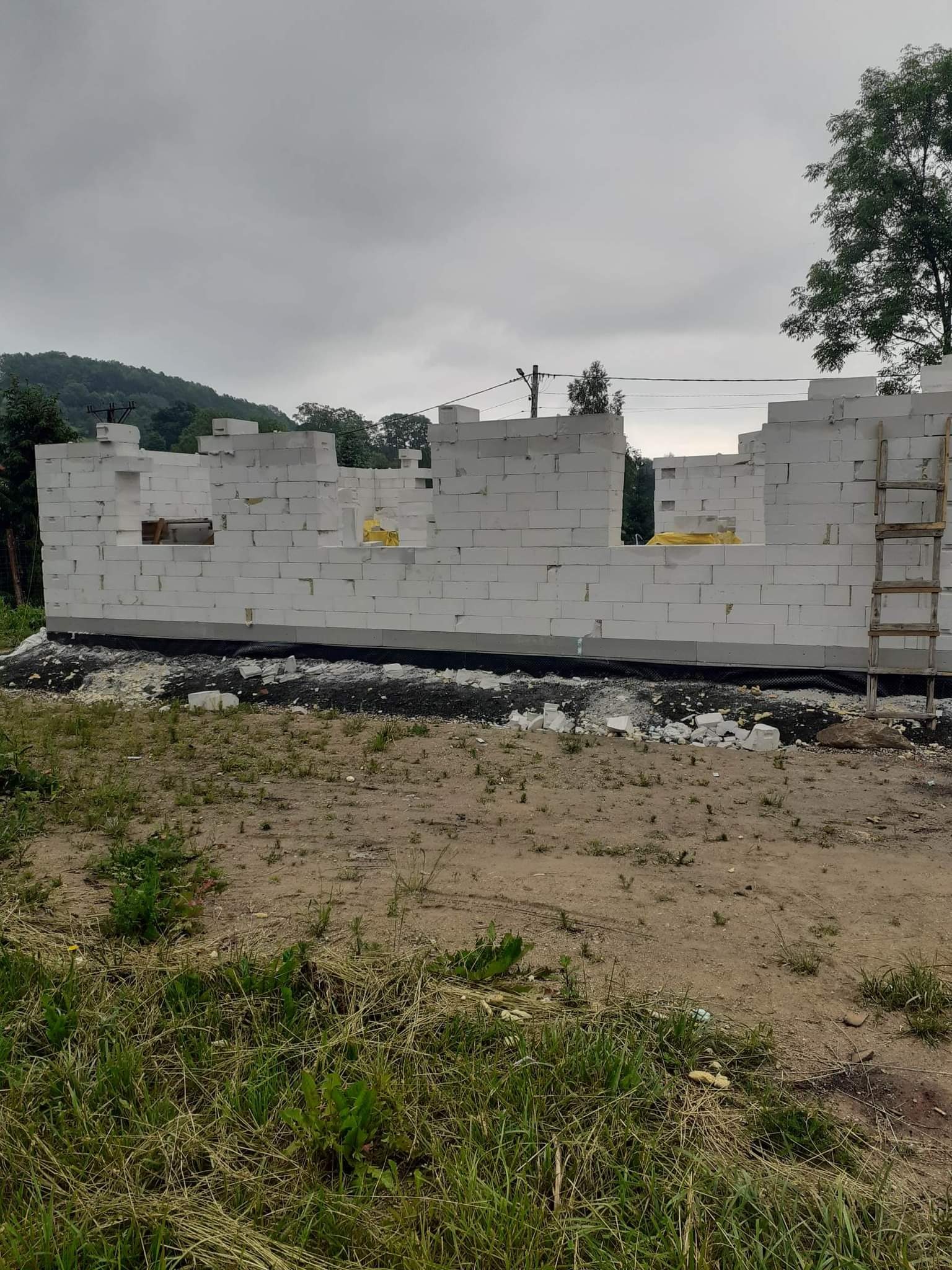 Wznoszone ściany domu z białych bloczków, widoczne z zewnątrz, z drewnianą drabiną opartą o ścianę, na tle pochmurnego nieba i zieleni wzgórz.